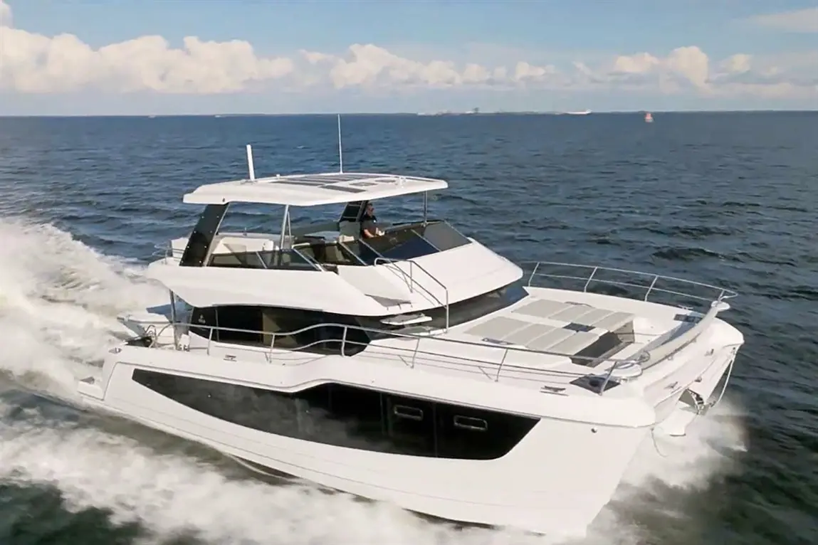 Luxury motor yacht cruising on blue waters with a captain at the helm, surrounded by clouds and distant boats.